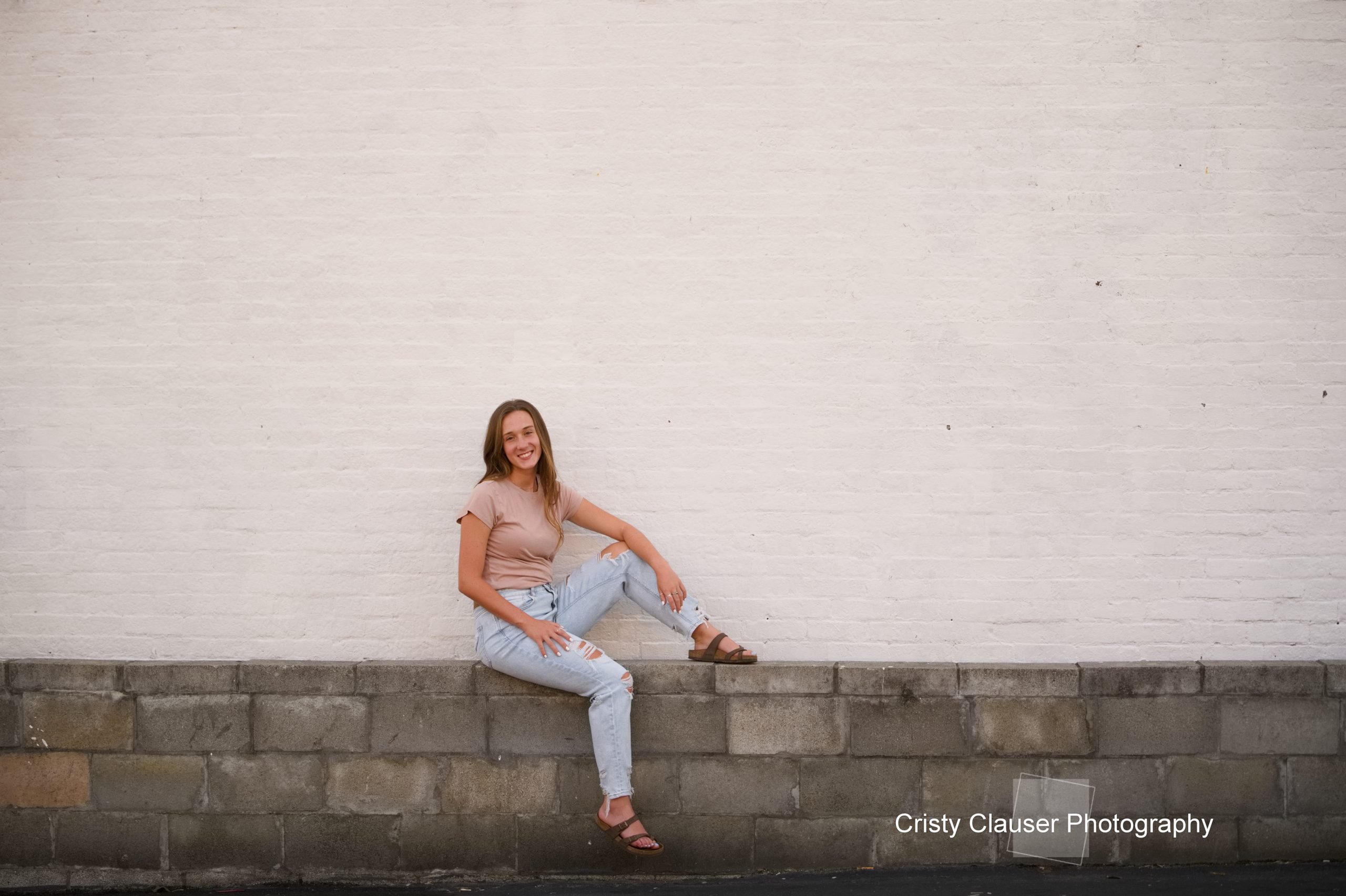 A woman seated on a stone wall, wearing a smile, with a backdrop of soothing neutral colors.