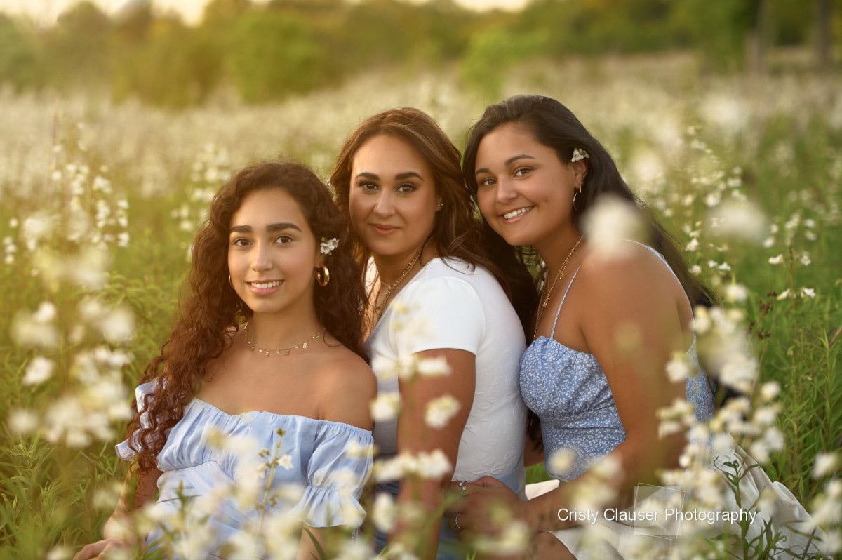 Three women stand in close-up smiling, surrounded by white flowers.
