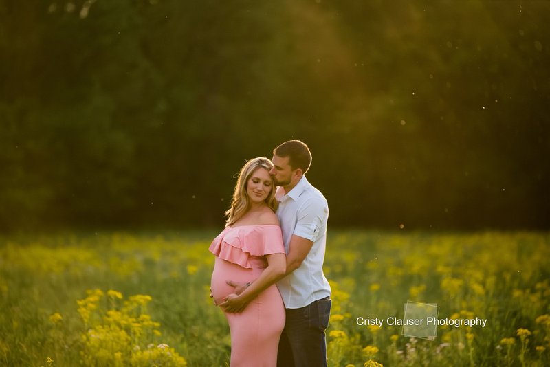 A pregnant mother closes her eyes and looks over her shoulder, while the father stands behind, holding her and placing a gentle kiss on her forehead, capturing a moment of shared love and anticipation.