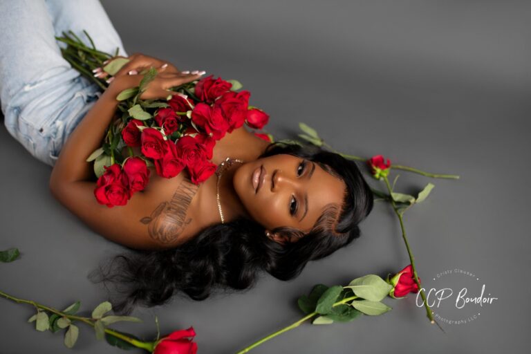 A woman lies on her back against a gray background, holding a bouquet of red roses close to her chest. She wears jeans, and her long black hair is spread out. Other roses are scattered around her.