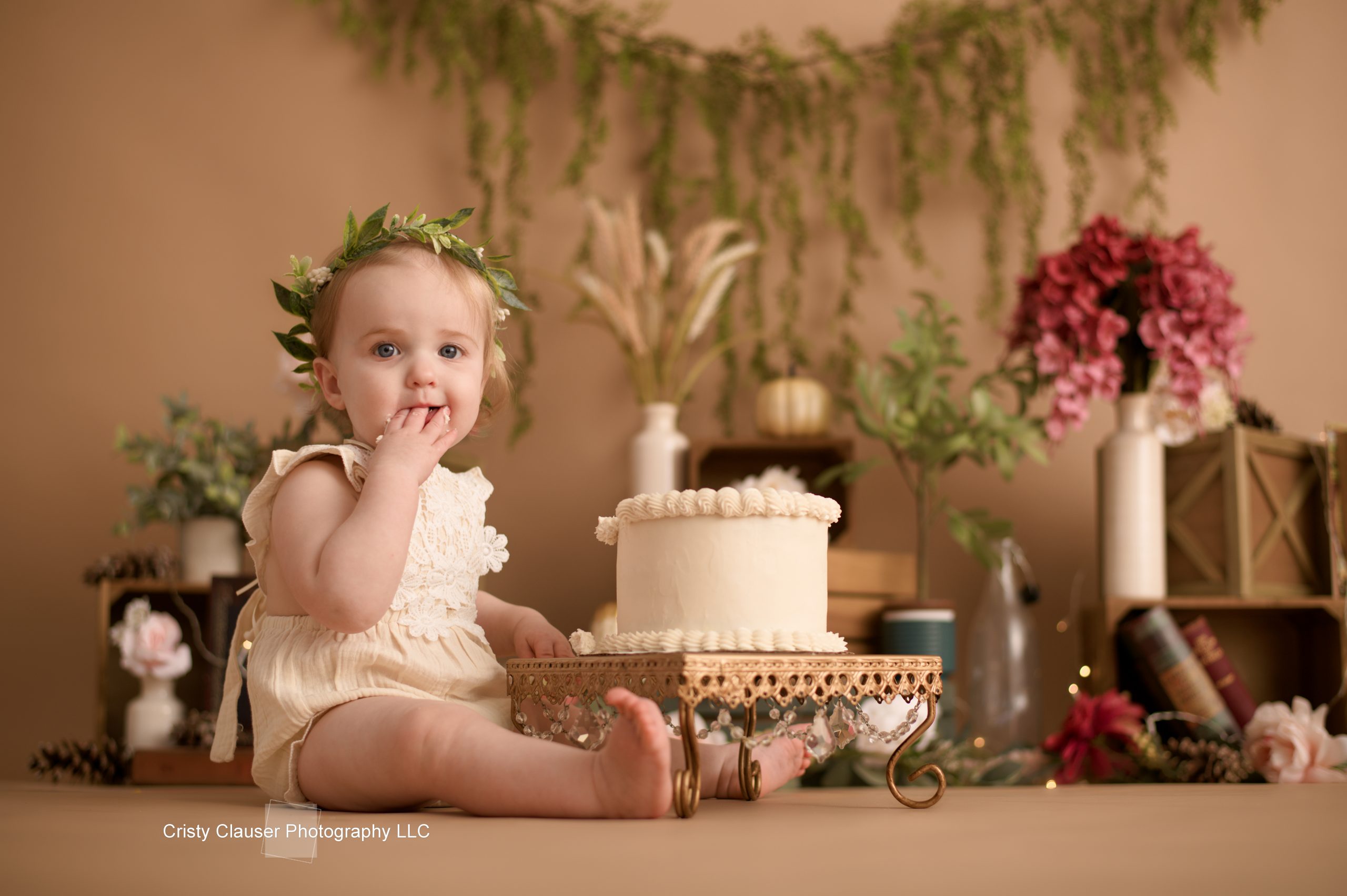 A baby in a white outfit with a floral headband sits on the floor next to a cream-colored cake on a stand. The background features greenery, flowers, and books.