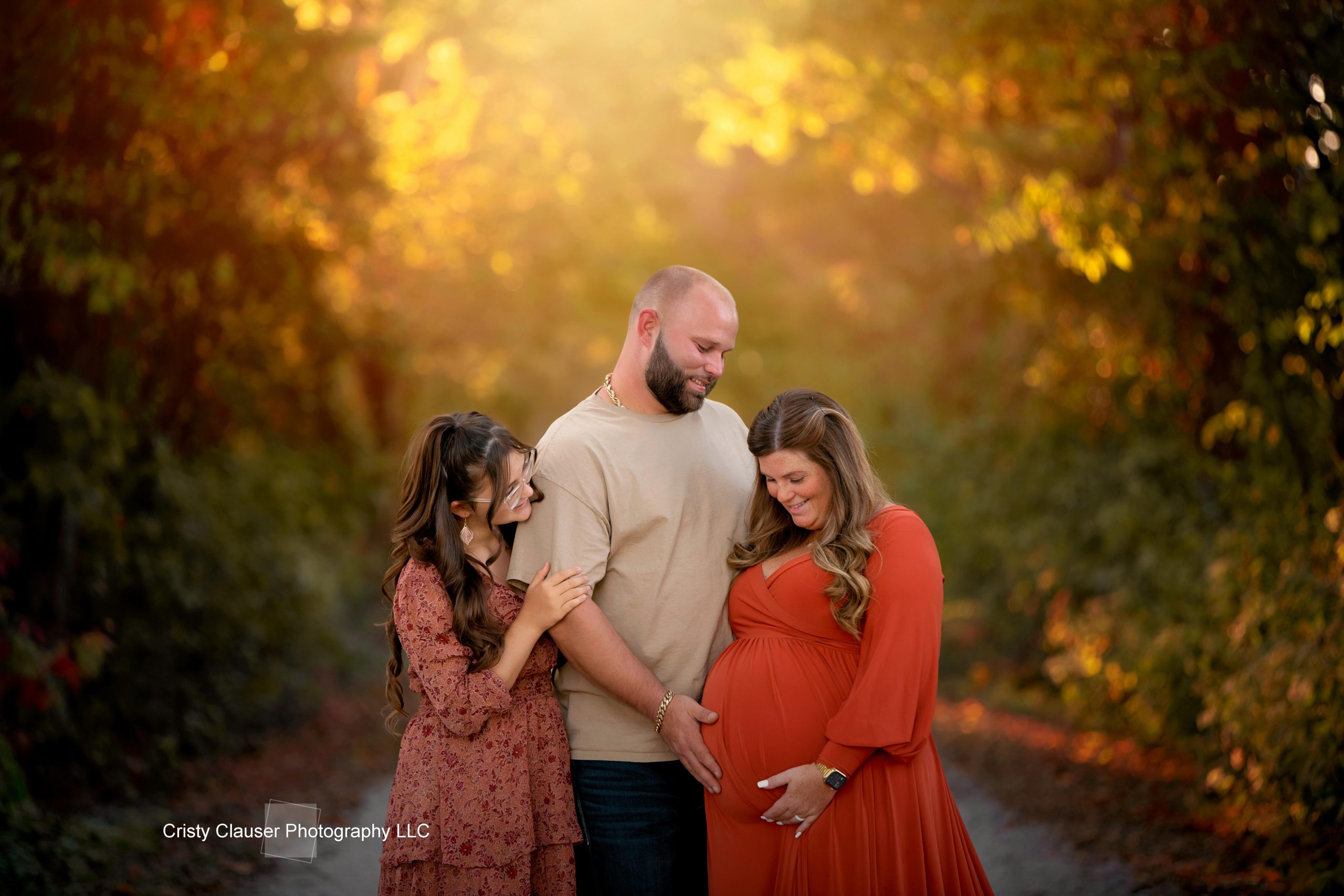 A couple stands in a sunlit path surrounded by autumn foliage. The woman holds her pregnant belly while her partner stands beside her. Another woman stands next to them, smiling and touching the pregnant woman's arm.