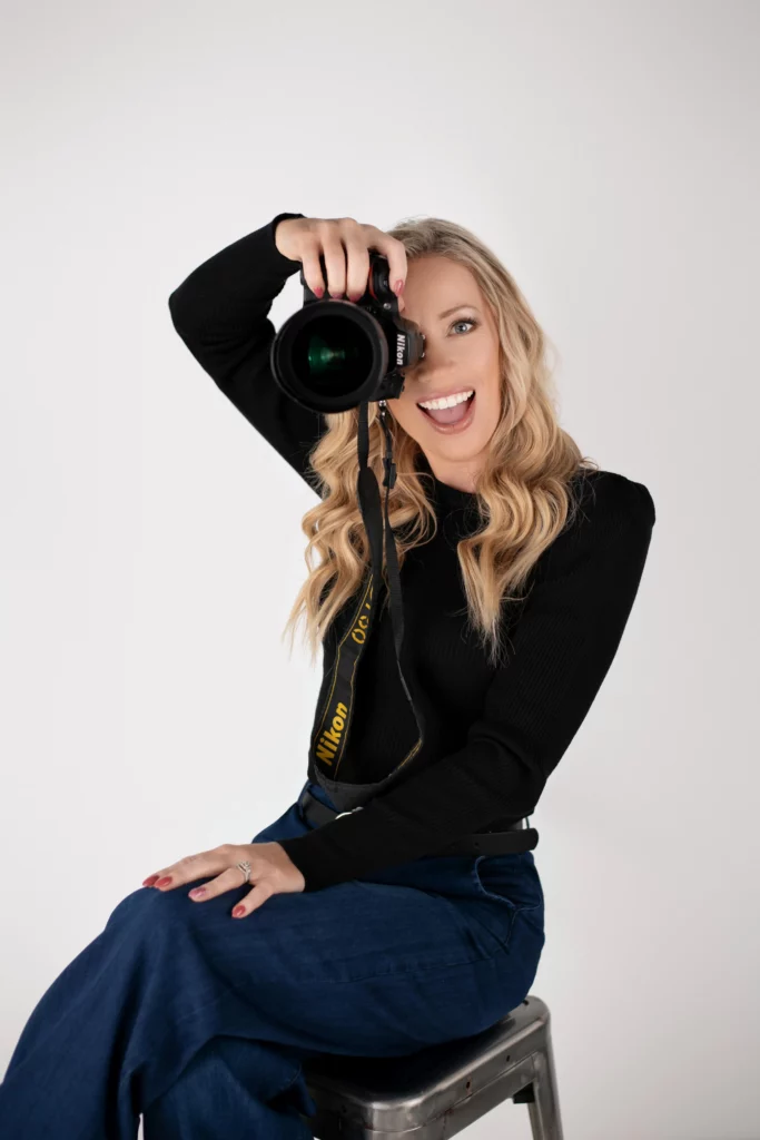 A smiling woman with long blonde hair, wearing a black top and blue jeans, sits on a stool holding a Nikon camera up to her face, posing playfully against a plain white background. Cristy Clauser Photography