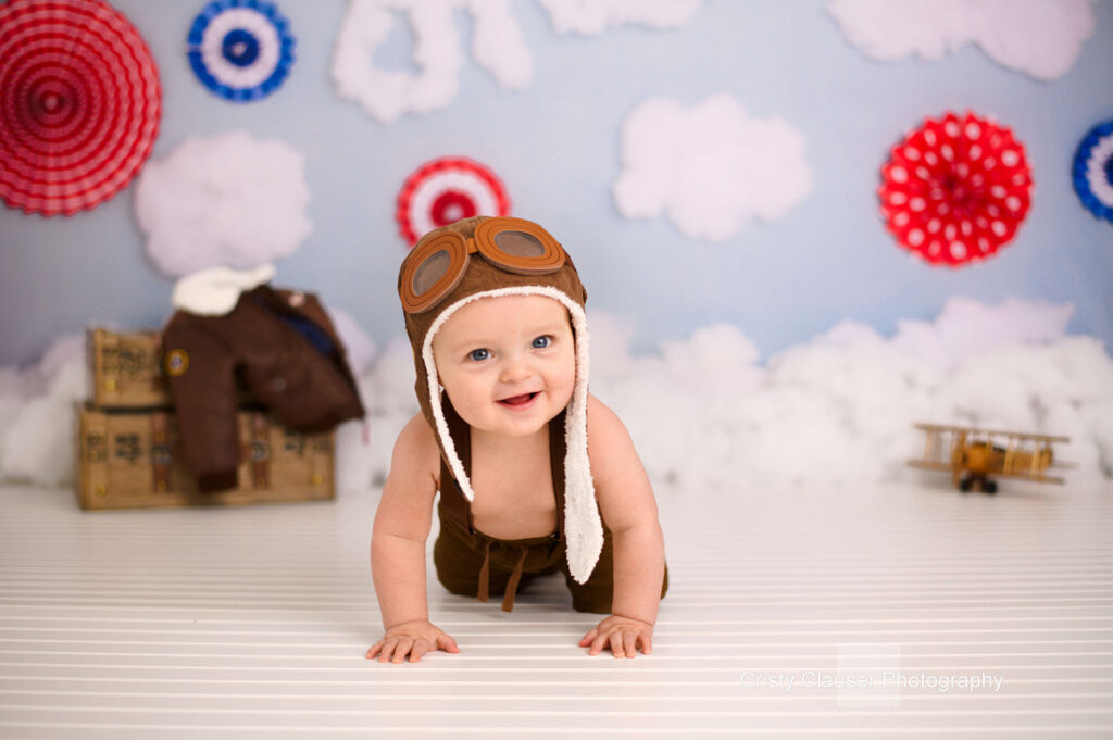 A baby wearing a brown aviator hat with goggles crawls on a white floor. In the background, there's a toy plane, a jacket on a box, and red, white, and blue decorations resembling clouds and target patterns.