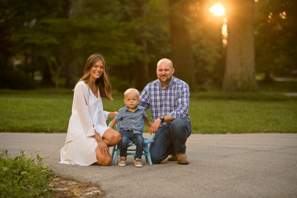 A woman, a man, and a young boy pose together outdoors on a paved path at sunset, with green grass and trees in the background. The child sits on a small blue chair, while the adults kneel beside him, smiling. Cristy Clauser Photography