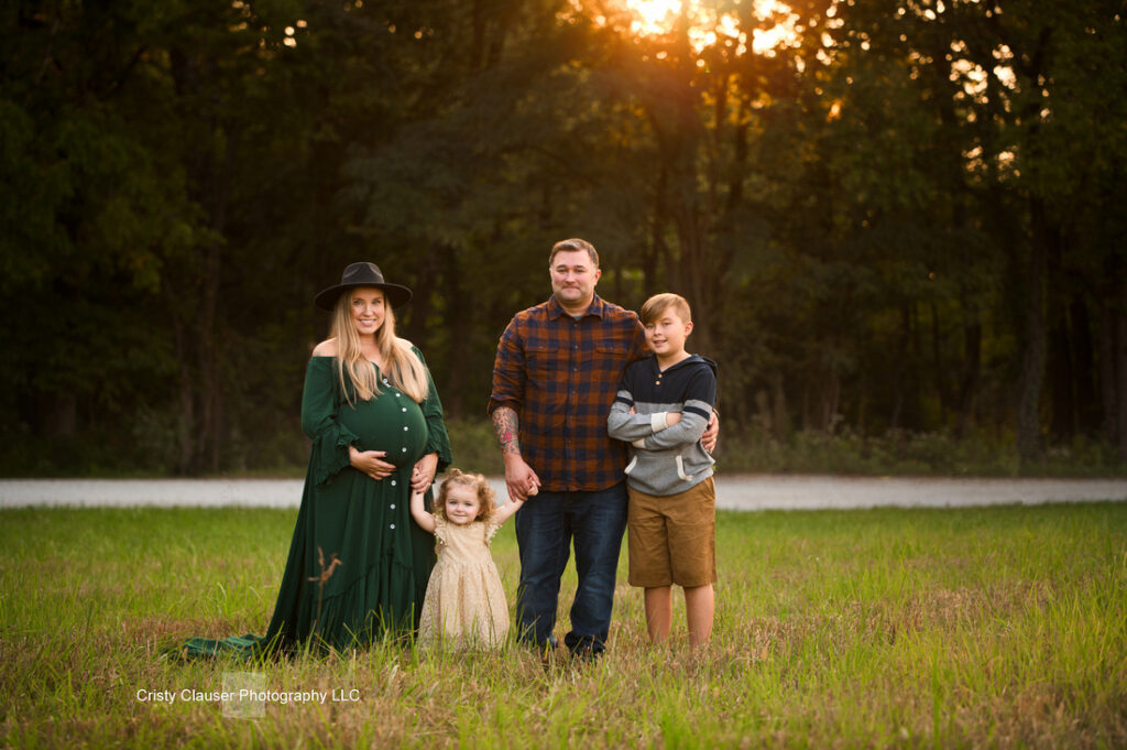 A pregnant woman, a man, a young girl, and a teenage boy stand together in a grassy field at sunset, with trees in the background. The woman wears a green dress and hat; all are smiling at the camera. Cristy Clauser Photography