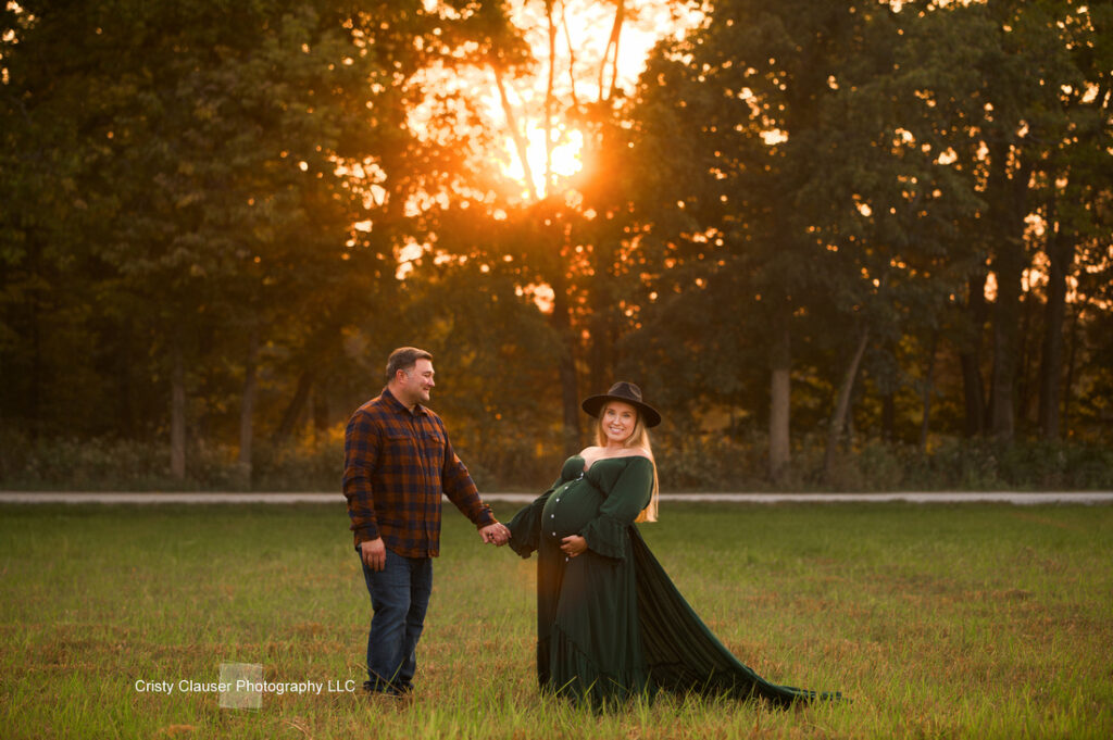 A couple stands in a grassy field at sunset. The man, in a plaid shirt and jeans, holds hands with a pregnant woman in a flowing green dress and black hat. Sunlight filters through trees in the background. Cristy Clauser Photography