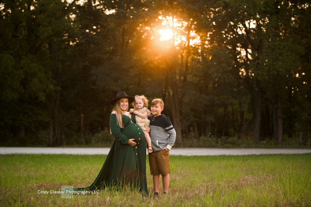 A pregnant woman in a long green dress and black hat stands in a grassy field at sunset, holding a small child. An older child stands beside her, and trees fill the background. Cristy Clauser Photography