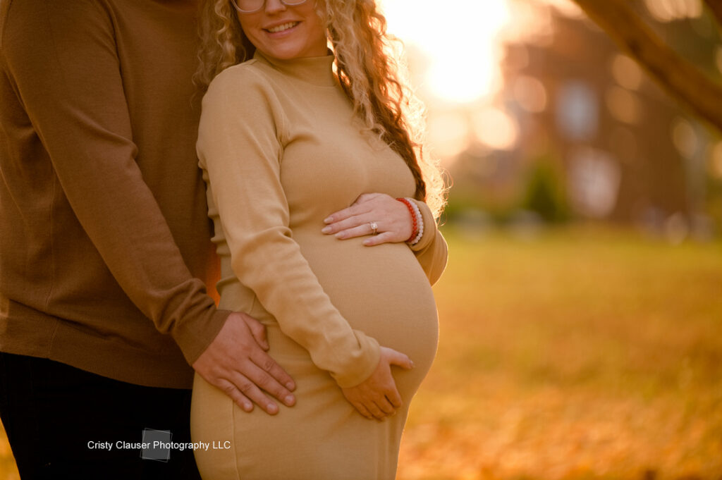 A pregnant woman in a tan dress stands outdoors at sunset, smiling as a partner stands beside her with hands gently placed on her baby bump. The background is softly blurred, with warm golden light and autumn foliage. Cristy Clauser Photography