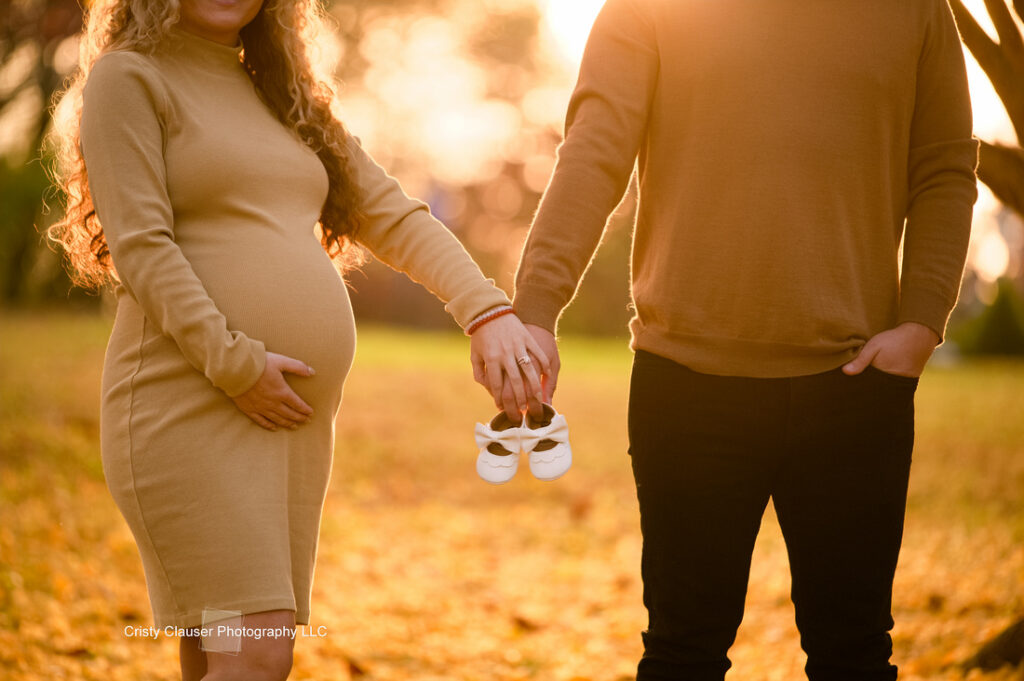 A pregnant woman and a man stand outside holding hands at sunset. The woman cradles her belly, and the man holds a pair of small white baby shoes, symbolizing their anticipation of a new baby. Cristy Clauser Photography