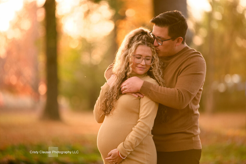 A man embraces a pregnant woman from behind in a sunlit park with autumn colors. Both wear tan sweaters and glasses, smiling warmly and looking content. Cristy Clauser Photography