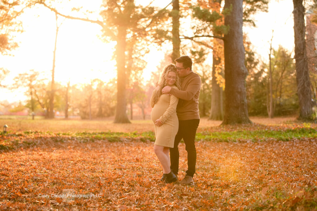 A couple poses in a sunlit park with autumn leaves on the ground. The woman, visibly pregnant and wearing a fitted dress, stands in front while the man embraces her from behind. Tall trees are in the background. Cristy Clauser Photography