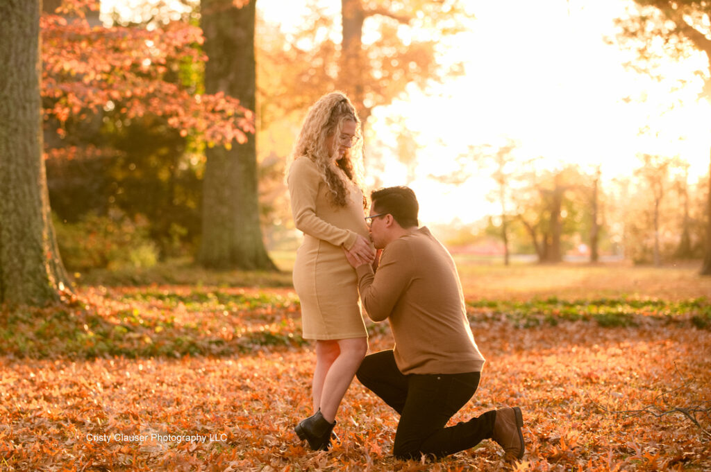 A man kneels on one knee, holding a woman's hand and kissing it, in a sunlit park with autumn leaves covering the ground. Both are dressed in warm tones, and golden sunlight streams through the trees around them. Cristy Clauser Photography