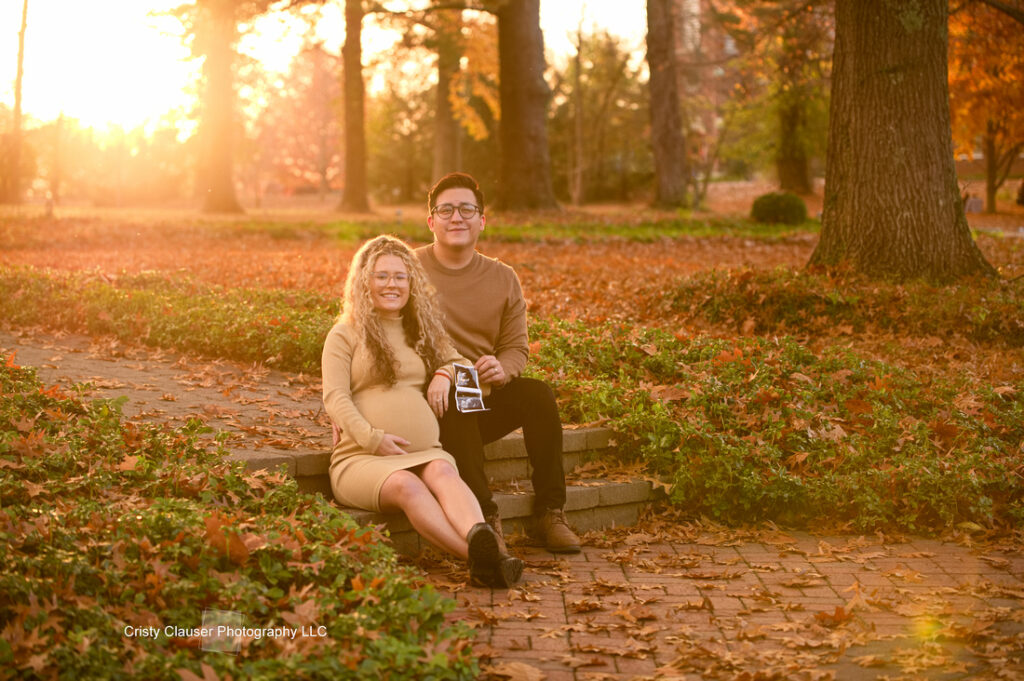 A couple sits on stone steps in a park covered with autumn leaves, smiling at the camera. The sun sets behind them, casting a warm golden light. The woman wears a tan dress; the man wears a tan sweater and holds an ultrasound photo. Cristy Clauser Photography