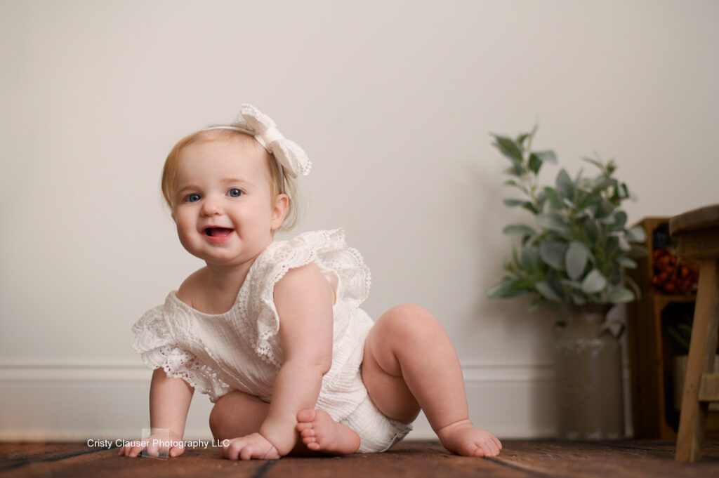 A baby wearing a white outfit with lace details sits on a wooden floor, smiling. A small bow adorns their hair. A plant in a pot and a woven basket on a stool are visible in the background against a plain wall.