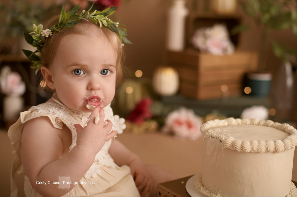 A toddler with a floral headband and cream-colored dress sits near a partially eaten cake, with frosting on her fingers. The background includes blurred flowers and decorations, creating a warm, festive atmosphere.