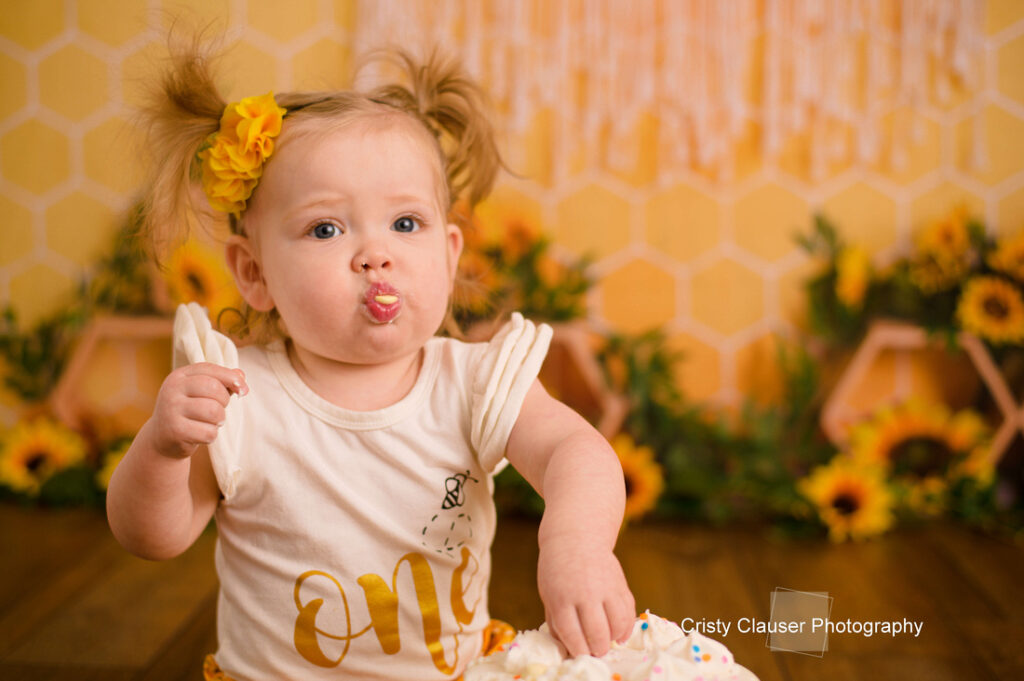 A toddler with blonde pigtails and a yellow flower hair clip wears a white "one" birthday shirt and eats cake. She sits in front of a sunflower-themed background and looks forward with cake on her face. Cristy Clauser Photography