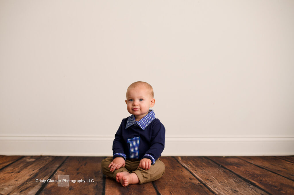A baby sits on a wooden floor against a plain white wall. The baby is dressed in a navy sweater with a blue and white checkered collar and khaki pants, looking towards the camera with a neutral expression.