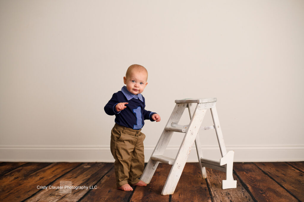 A baby stands next to a white wooden step stool on a wooden floor. The baby is wearing a navy blue shirt and khaki pants, with bare feet. A white number "1" is on the stool. The background is a plain white wall.
