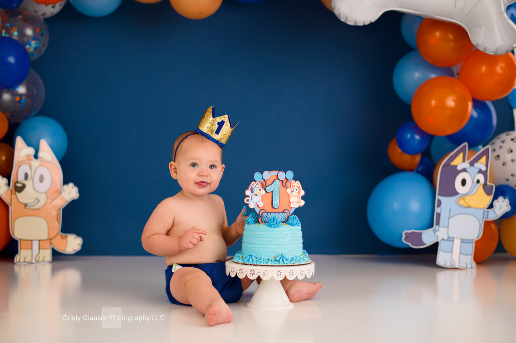 A smiling baby wearing a crown sits next to a blue birthday cake topped with a number one. The background features colorful balloons and cartoon cutouts.