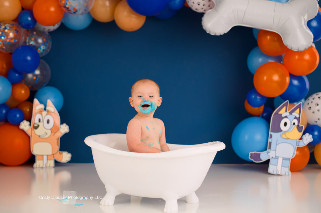 A smiling baby sits in a small white tub with blue frosting on their face. The background features colorful balloons and cutouts of animated dog characters.
