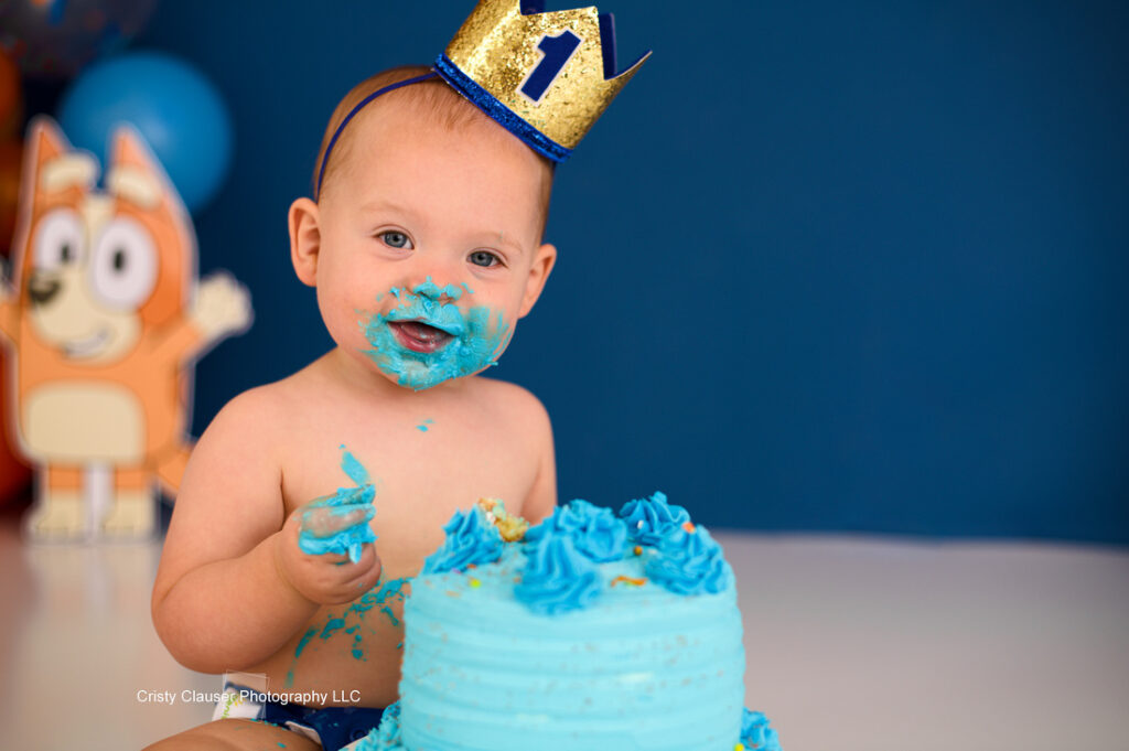 A baby wearing a gold crown with the number one on it is covered in blue frosting from a birthday cake. The cake is blue with swirled decorations. A cartoon figure is in the background against a blue wall.