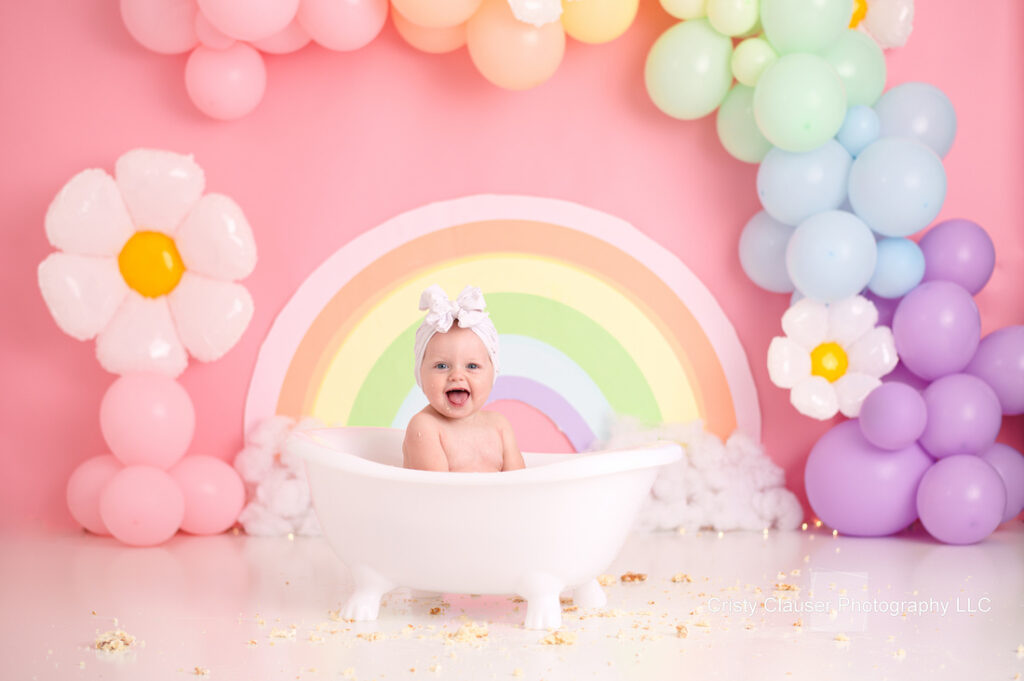 A baby sits joyfully in a white tub, surrounded by pastel-colored balloon arrangements resembling a rainbow and clouds. Large balloon flowers accent the setup against a pink backdrop. The scene is playful and whimsical.