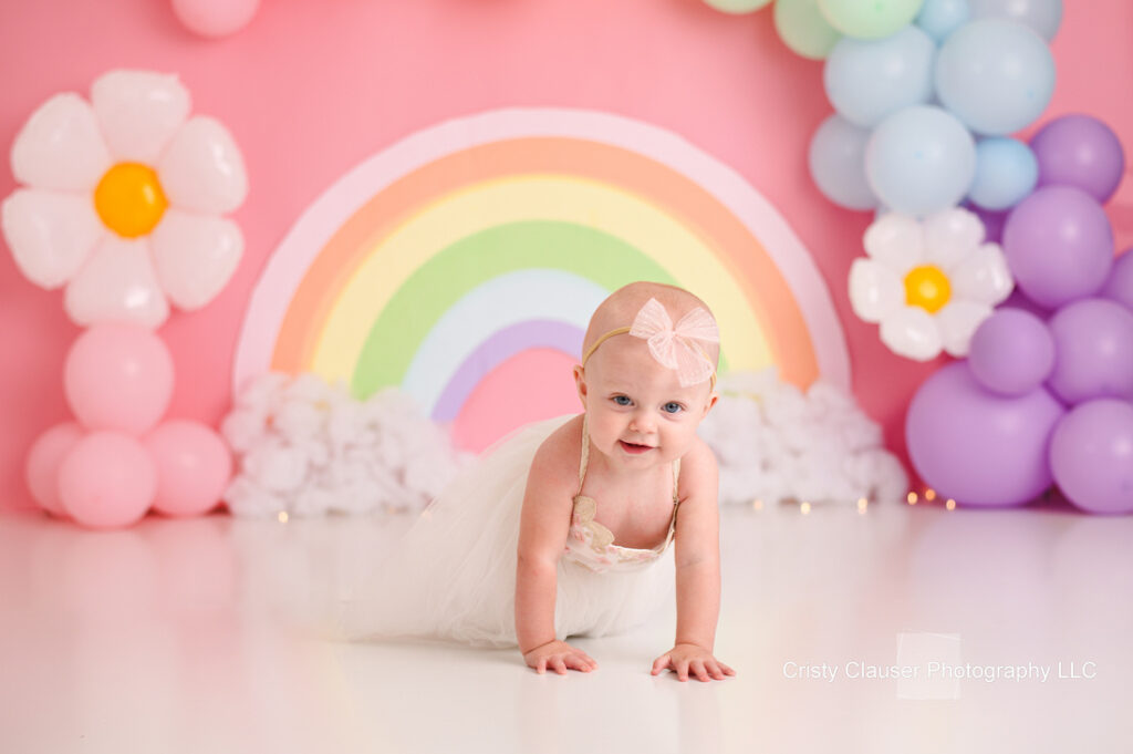 A baby dressed in a white outfit and pink bow headband crawls on a white floor. The background features a colorful rainbow mural and pastel balloon decorations, including large white flowers with yellow centers.