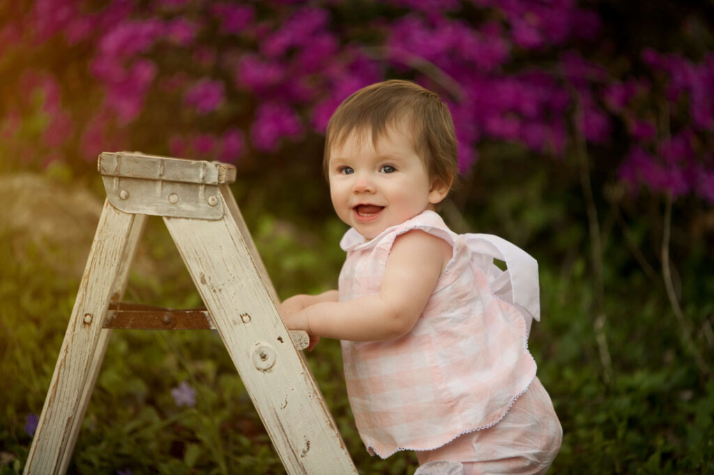 A smiling baby in a pink plaid outfit stands holding a small white wooden ladder, with bright purple flowers and greenery in the blurred background. Cristy Clauser Photography