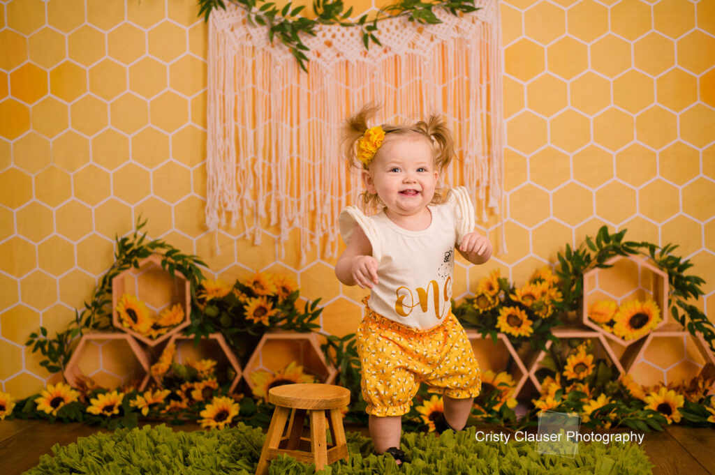A toddler with pigtails smiles joyfully while standing near a small wooden stool. The child wears a white shirt and yellow pants, with a matching hair bow. The background features a honeycomb pattern, sunflowers, and a woven decoration.