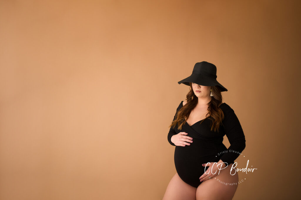 A pregnant woman in a black outfit and wide-brimmed hat poses against a plain tan background, holding her belly with one hand and her thigh with the other. The photo is watermarked “JCP Boudoir Photography LLC.”. Cristy Clauser Photography