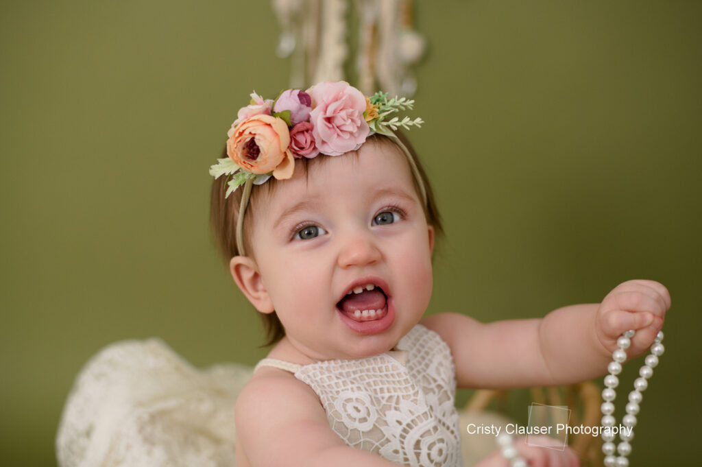 A baby wearing a lace outfit and a floral headband smiles while holding a string of pearls. The background is a soft green.© Cristy Clauser Photography.