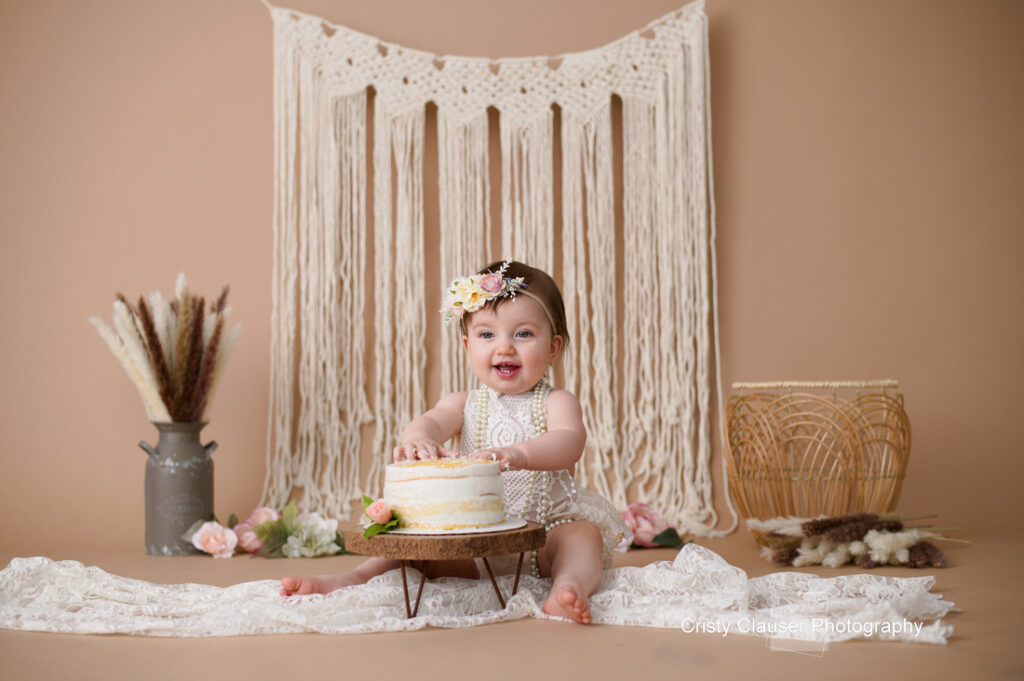 A joyful baby sits in front of a small cake, wearing a floral headband and lace outfit. The background features a macramé wall hanging, dried flowers in a vase, and a wicker basket. Cakesmash setting with a beige color scheme.
