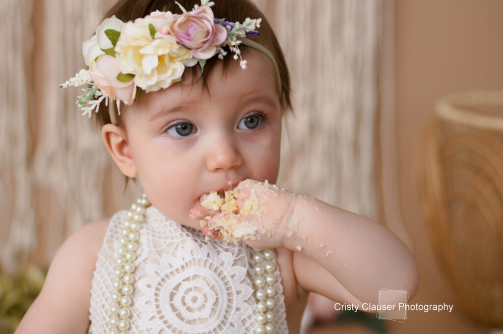 A baby with a flower headband and pearl necklace is eating cake. Her hand is covered in cake crumbs as she takes a bite. She is wearing a white lace dress. The setting is soft and neutral with hints of floral decor.