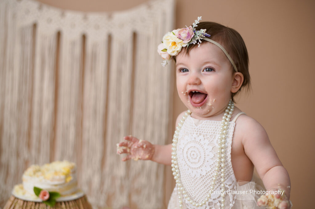 A joyful baby with cake on her face and hands wears a floral headband and a pearl necklace. She sits in front of a partially eaten cake and a macrame backdrop.