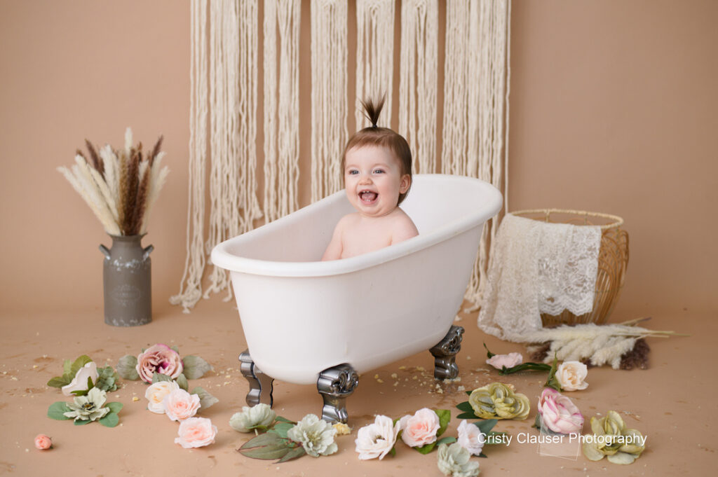 A baby is smiling while sitting in a small white bathtub placed on a floor decorated with pastel flowers. The backdrop features hanging macrame and arranged pampas grass. The setting has a warm, cozy feel.