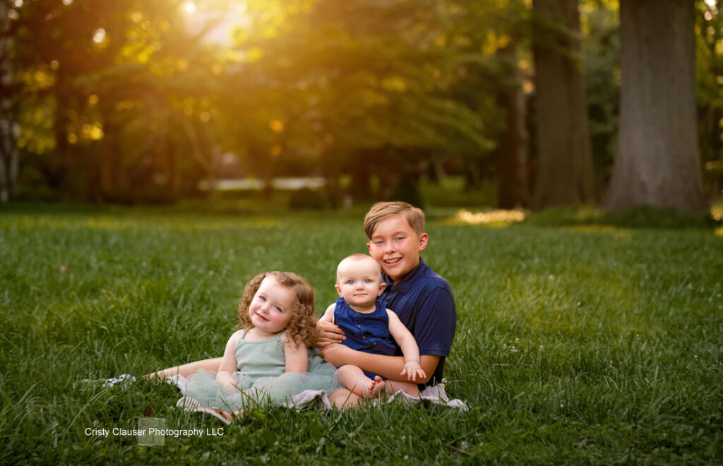 Three young children sit together on green grass in a sunlit park, smiling at the camera. Tall trees and soft sunlight create a warm, peaceful background. Cristy Clauser Photography