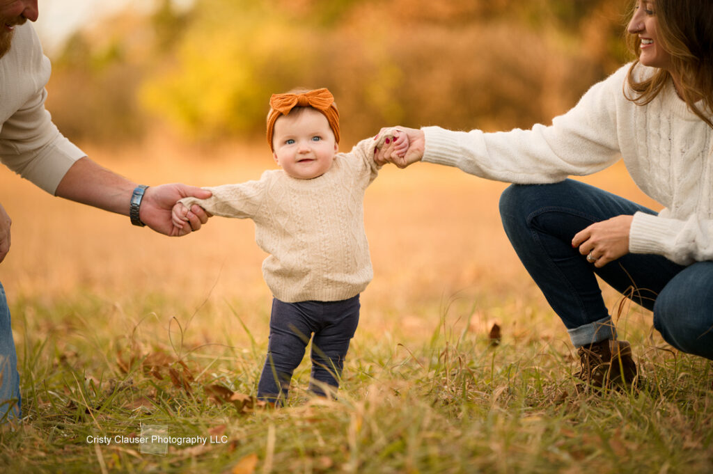 A smiling baby wearing a knit sweater, blue pants, and an orange bow stands in a field holding hands with two adults, both dressed in cozy, neutral sweaters. Autumn foliage is visible in the background. Cristy Clauser Photography