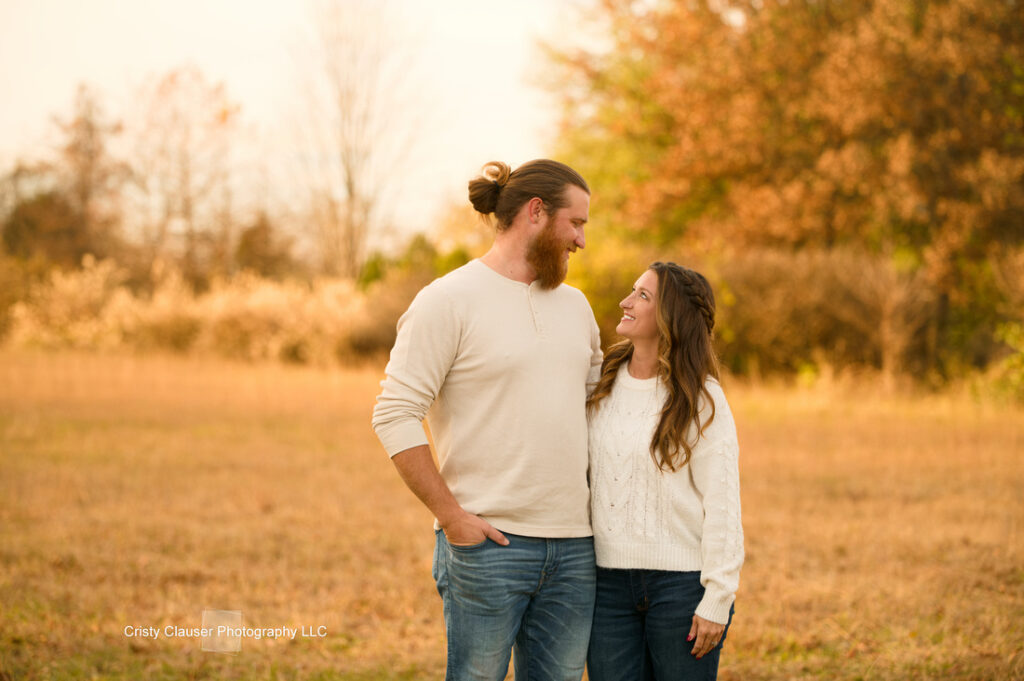 A man and woman stand outdoors in a golden field, smiling at each other. Both wear casual light sweaters and jeans, with autumn trees blurred in the background. The scene feels warm and relaxed. Cristy Clauser Photography