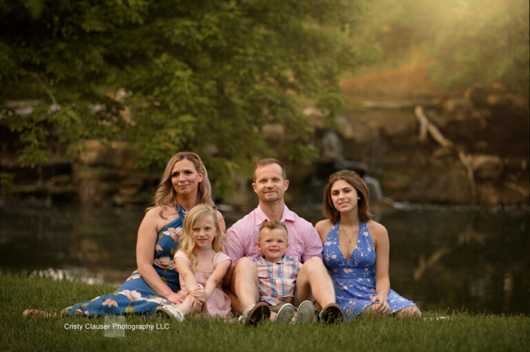 A family of five sits on grass by a pond, smiling at the camera. The parents are in the center with two daughters and a son around them. Trees and sunlight create a warm, natural background. Cristy Clauser Photography