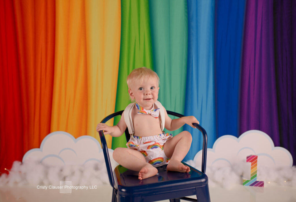 A smiling baby sits on a dark blue chair in front of a rainbow-colored backdrop, which includes clouds and the number "1" in rainbow colors. The baby is wearing a rainbow-patterned outfit and looks cheerful.