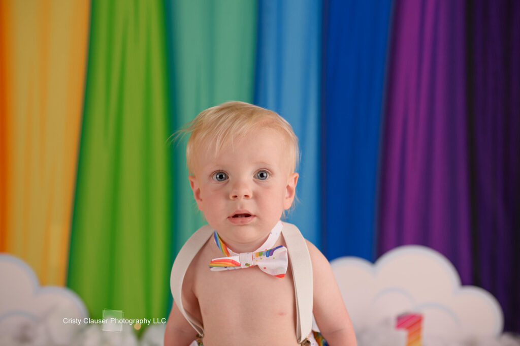 A baby with blonde hair is wearing a colorful bow tie and suspenders. They are sitting in front of a rainbow-colored backdrop with clouds at the bottom.