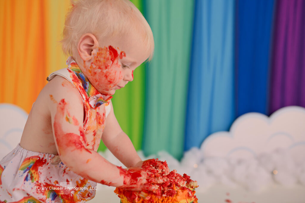 A toddler with a rainbow-patterned outfit is joyfully playing with a cake, covered in colorful frosting. The background features curtains in various rainbow colors and a cloud decoration.