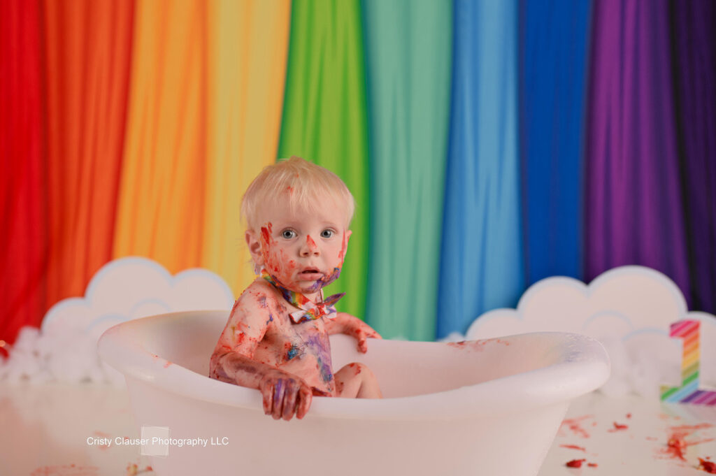 A baby with blonde hair sits in a white tub, covered in colorful paint. The backdrop features a vibrant rainbow curtain with cloud cutouts at the bottom. The baby's expression is curious and slightly surprised.