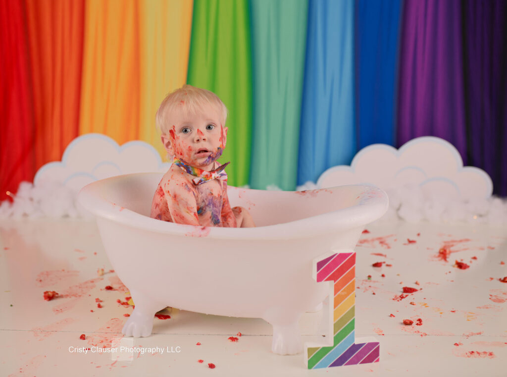 A baby covered in colorful cake sits in a white bathtub prop with a rainbow backdrop. Frosting and cake pieces are scattered around. There's a large number one in the foreground, signifying a first birthday celebration.