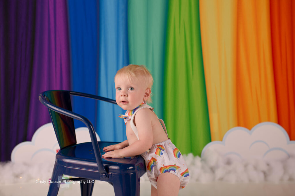 A toddler wearing a rainbow-patterned outfit stands next to a dark chair in a room with a vibrant rainbow backdrop and cloud decorations.