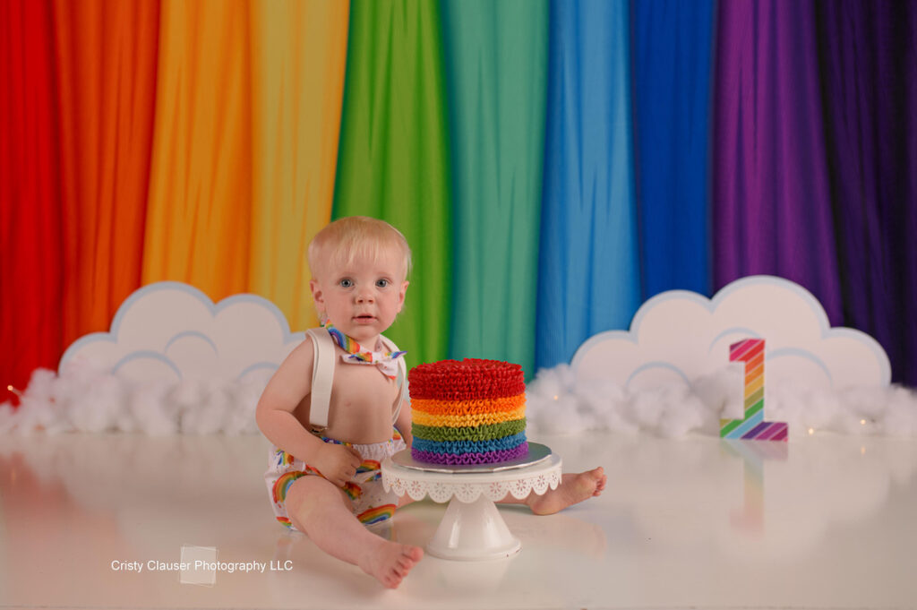 A baby sits on a reflective floor in front of a rainbow backdrop with a giant number one. The baby wears rainbow-themed suspenders and shorts, and a rainbow cake is placed on a stand beside them.