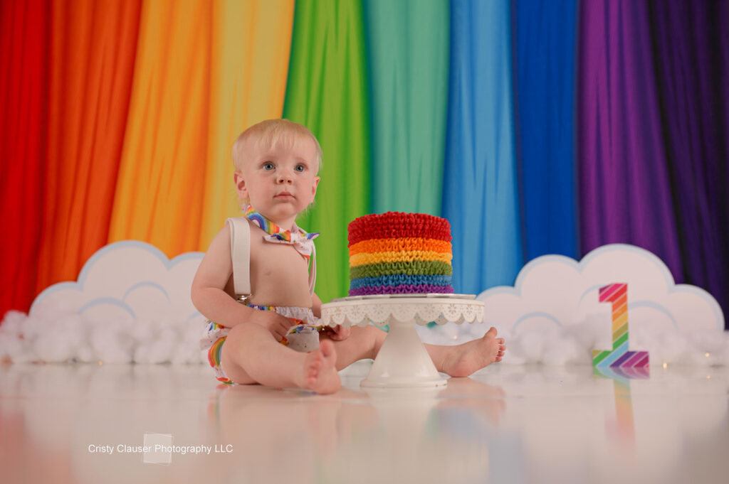 A toddler sits on the floor in front of a rainbow backdrop, wearing suspenders and a bow tie. In front of them is a small rainbow cake on a cake stand. A number "1" decorated with rainbow colors is nearby.