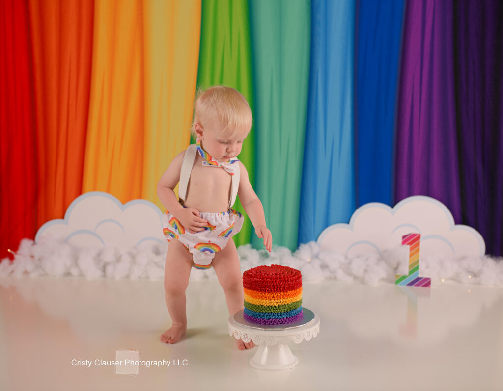 A toddler in rainbow-themed attire stands in front of a rainbow backdrop with clouds. The child reaches toward a colorful rainbow cake on a white stand. A number "1" is displayed nearby, indicating a first birthday celebration.