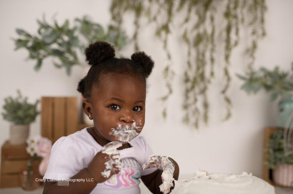A toddler with two buns in her hair looks at the camera with cake on her face and hands. She is wearing a white shirt, and there is foliage and decorations in the background.